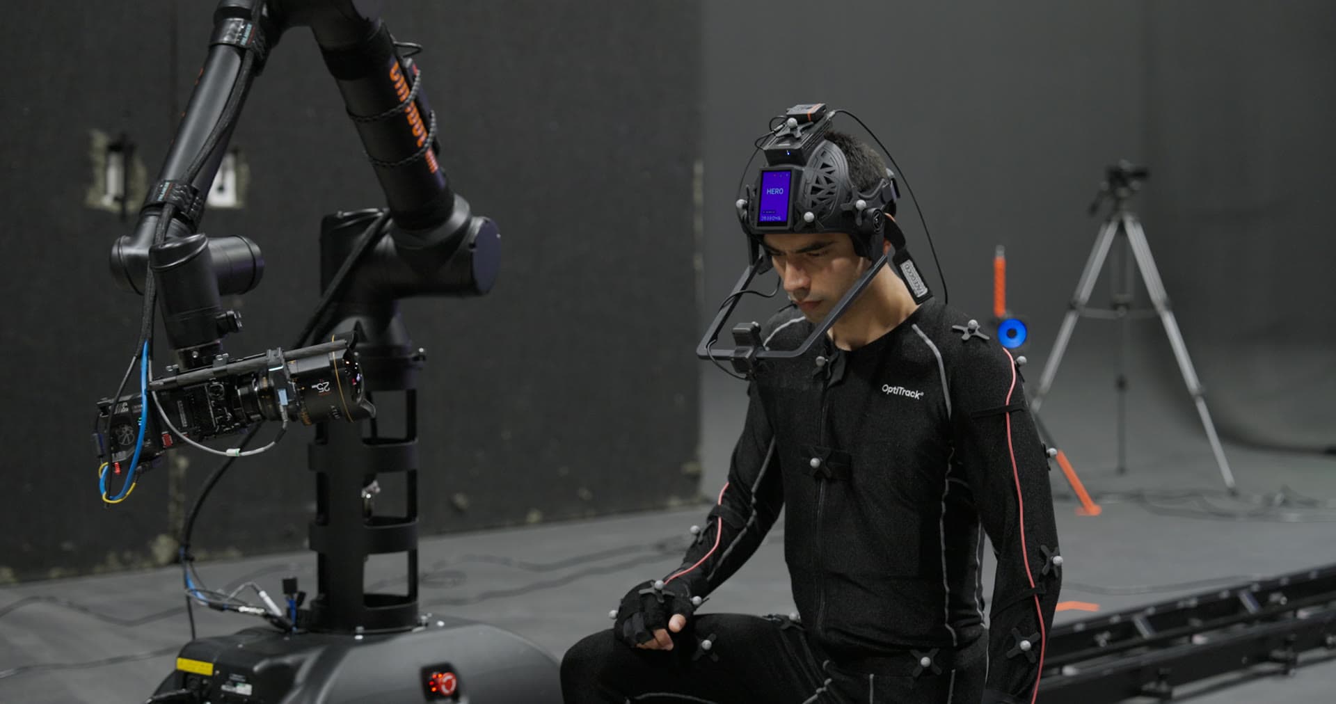 Performer in an OptiTrack motion capture suit wearing a FACEGOOD SMART P3 head-mounted camera, timecode on the helmet touchscreen, beside a robotic camera arm at the Form Volumes studio